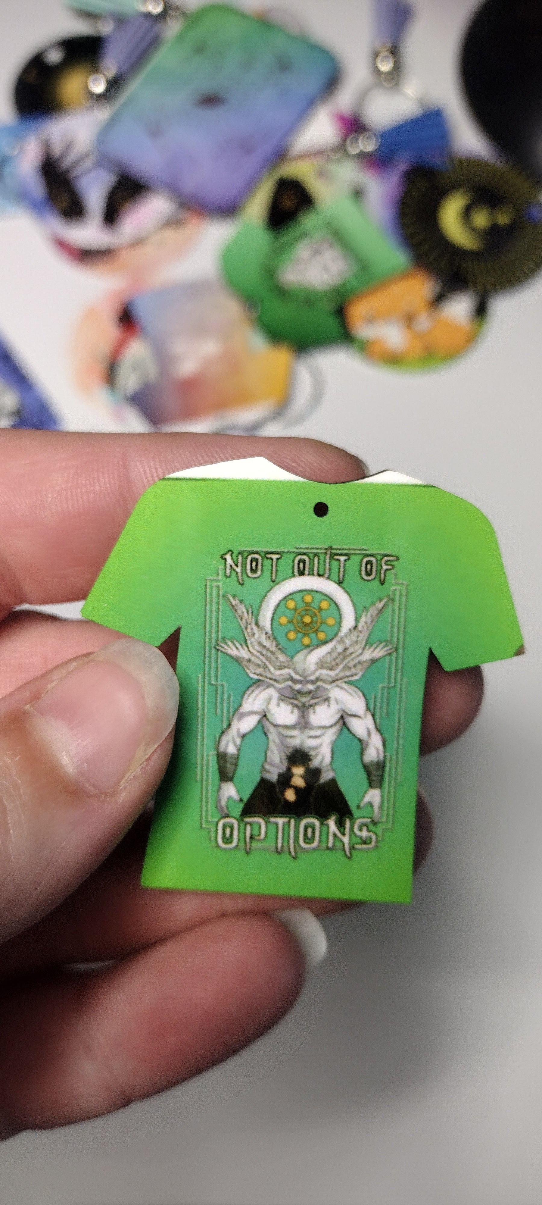 closeup of a tshirt shaped keychain that has an image of an anime character Megumi in front of Mahoraga. The keychain is imperfect, with a white line on the top. 